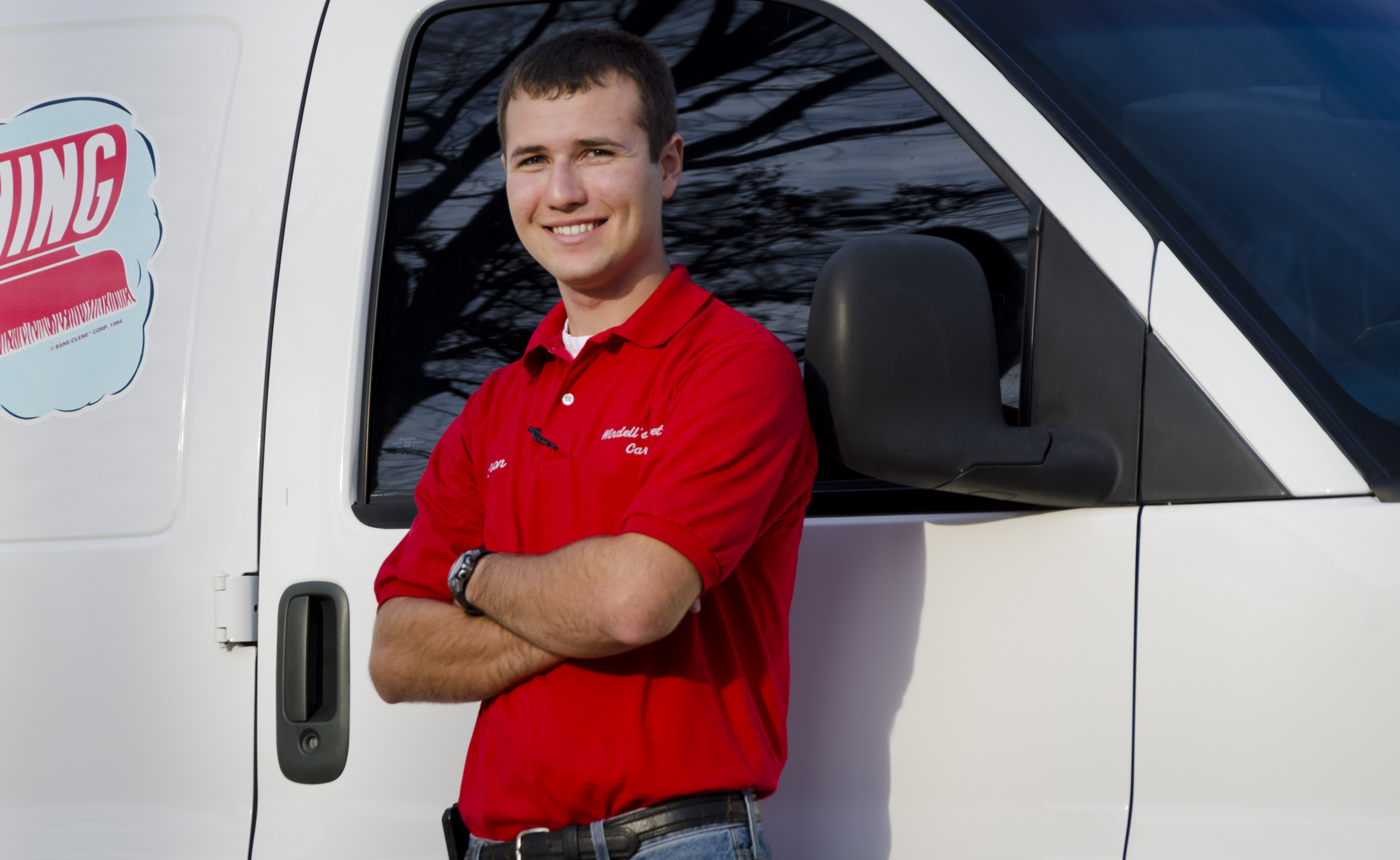 A person in a red polo shirt and jeans stands with arms crossed and smiles in front of a white van featuring a partially visible best carpet cleaning business logo.
