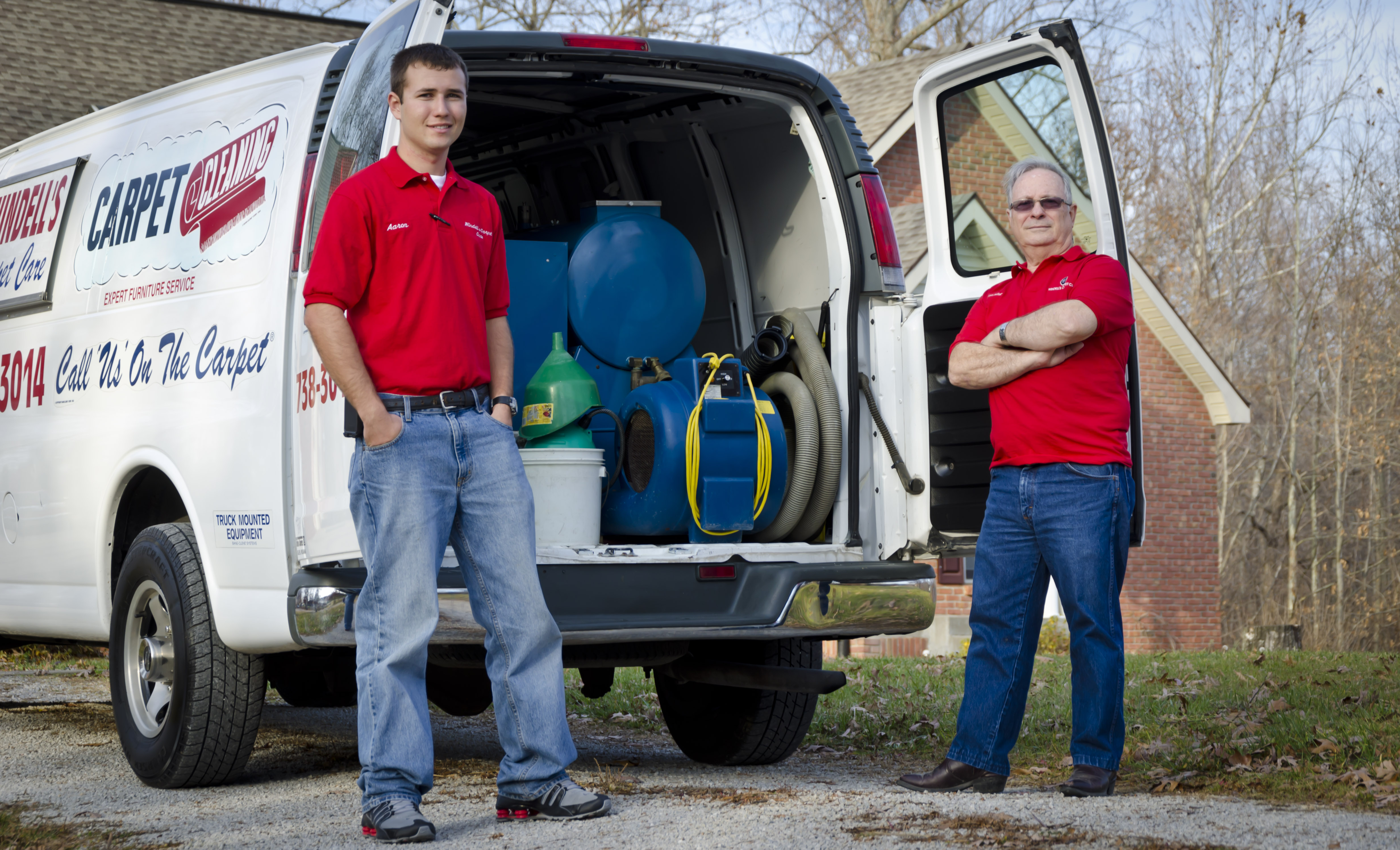 Two men in red shirts and jeans stand beside an open white van, filled with the best carpet cleaning equipment, parked on a driveway near a brick house and trees.