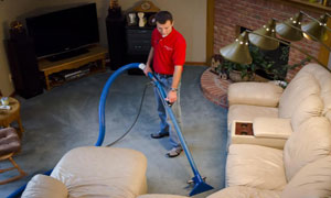 A man in a red shirt uses professional equipment to clean the carpet in a living room with cream-colored sofas, a TV, and a brick fireplace—perfect for those seeking expert carpet cleaning in Floyd Knobs or southern Indiana.