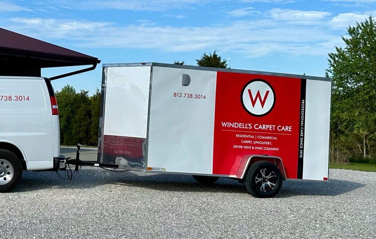 A white trailer with a red and black Windells Carpet Care logo and contact info is attached to a white van, parked on gravel with trees and blue sky in the background—your trusted choice for carpet cleaning in Floyd Knobs and Lanesville.