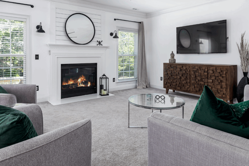 A portrait of a modern, clean living room with a fireplace and two windows on the far wall, a TV and wooden cabinet on the side wall, and a round coffee table on a clean, gray carpet.