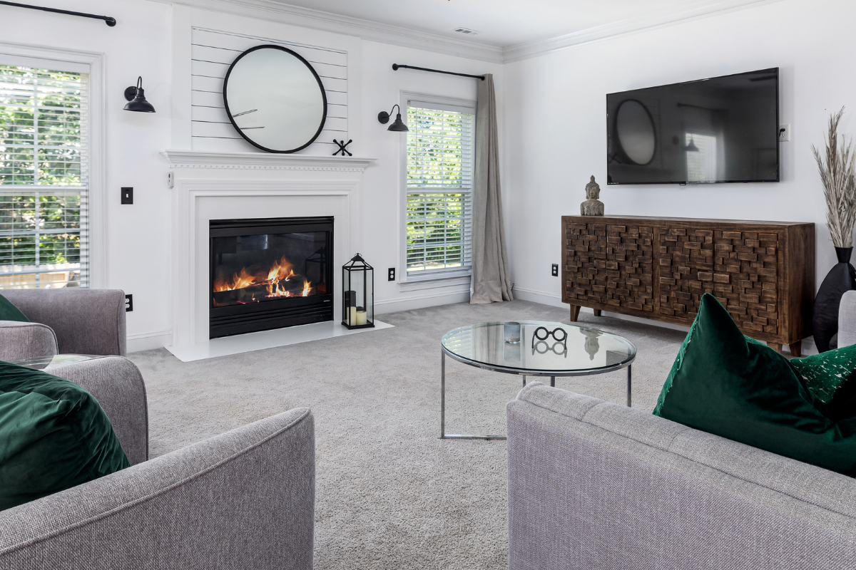 A portrait of a modern, clean living room with a fireplace and two windows on the far wall, a TV and wooden cabinet on the side wall, and a round coffee table on a clean, gray carpet.