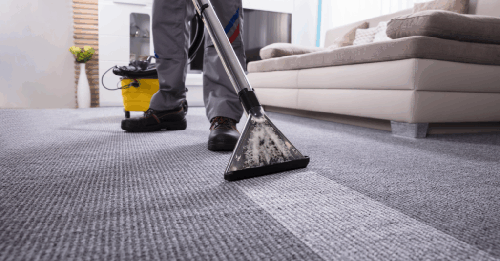 A person is cleaning a gray carpet in a living room with a professional carpet cleaning machine, reflecting the expertise of professional carpet cleaners. The scene includes a beige sofa, a yellow cleaning bucket, and sunlight streaming through a window.